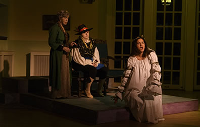 Production photo by Genneth Garrett of Ophelila in a white nightgown sitting on the edge of a platform with Osric sitting on a chair and a waiting woman standing by behind Ophelia. 