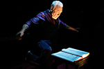 Patrick Page in dark purple shirt and black vest and pants, his face glowing in a spotlight, has his arms outstretched as he leans over an open book on the table. Photo by Julieta Cervantes.