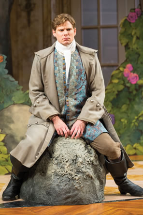 Dorante straddles a gray boulder, whearing long gray riding coat over floral-patterend waistcoat, high-collared white shirt, brown britches, and brown leather boots.