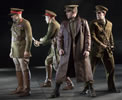 Othello and others stand in military uniforms with olive green service jackets, khaki jodhurs, brown boots, billed caps with red bands around them; two of the soldiers have their swords drawn; Iago stands in front, legs wide apart, in shin-length brown leather trench coat.
