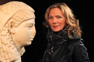 White bust of Cleopatra profile with sculpted head dress from the left looking right, and Cattrell in black leather coat and leopard print scarf in the background looking at the camera