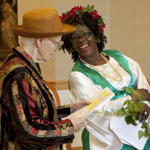 Old woman in multi-colored and black striped jacket, orange straw hat with script in hand next to a black woman with white blouse, bright green vest, and flowers in her hair