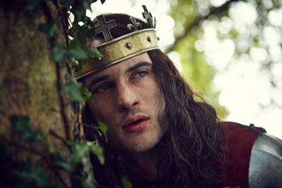 Henry VI, with a cross-topped gold headband as a crown, long hair, burgundy cloak over his armor, and bloodshot eyes, leans against an ivy-find tree trunck.