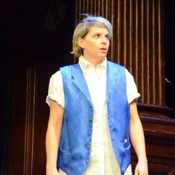 Julia in white work shirt, satin blue vest with subtle diamond pattern and lapels, and blue hair stands and looks to the side.
