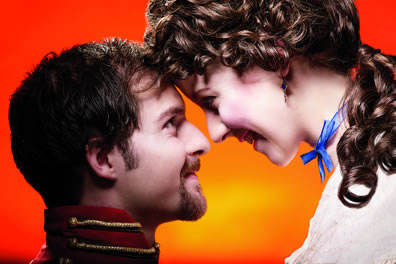 Closeup of Benedick and Beatrice smiling  at each other with foreheads touching against a bright orange background.