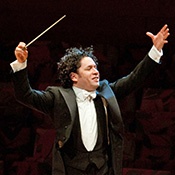 Dudamel, in tuxedo with black vest and white bowtie, hands upraise, baton in right hand, and hair flying.