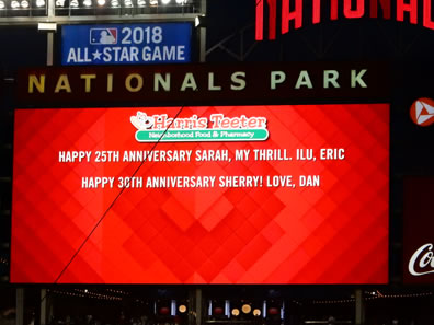 Nationals Park scoreboard, red background, Harris-Teeter Neighborhood Food & Pharmacy logo on top, two lines of type in white: "Happy 25th Anniversary Sarah, my thrill. ILU, Eric" and "Happy 30th Anniversary Sherry! Love, Dan."