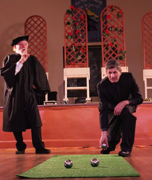 Holofernes in an academic gown and morter board with a finger up to his lips and his left hand on his hips talking as Nathanial kneels on one knee to cast a metal ball down an Astroturf rug where two other balls already sit: in the background, on a stage  are trellises of roses on wheeled countertops