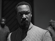 Corey Hawken in tunic and dark shadow on his face with a couple of other men in the background against a wall (black and white photograph).