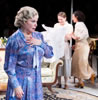 Fanny in blue floral print dress has her left hand at her chest, while in the background, David in light blue shirt and tan slacks sits on the couch, one foot crossed under the other leg, watching Marthe in a white blouse and ankle-lengh tan-striped skirt opening shopping bags with Babette.