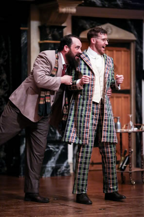 Toby in country light gray jacket and dark gray slacks, a scarf hanging from his neck, urges Aguecheek from behind, who is wearing a green and purple plaid zoot suit with white vest, his hands held up in an eager clinch: a drinks cart is in the background.