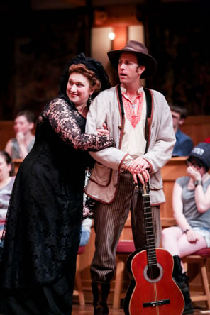 Olivia in black dress with lace sleeves and black hat leans against Feste with her arms in his as he stand wearing a hobo hat, simple jacket, Illyrian folk shirt and striped pants, his guitar standing on the floor with his hands folded on the key end. Audience on gallants' stools in background.