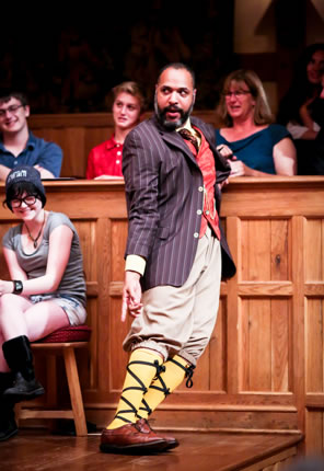 Leaning sideways against the theater wall, Malvolio in blue striped suit jacket, red patterend vest, red tie, tan knee pants, poins down to his yellow-stockinged, cross-gartered shins. Laughing girl sitting on gallant stool next to him, other audience in the background.