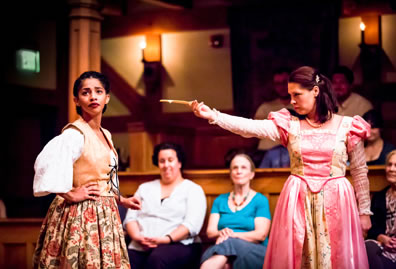 Julia in a pink Elizabethan dress with puffy shoulders, lace sleeves, and embroidered trim, holds a letter out to Lucetta, wearing a floral print pleated dress, brown leather vest tied at the front, and white blouse; she has her hands on her hips and facing away from the frowning Julia.