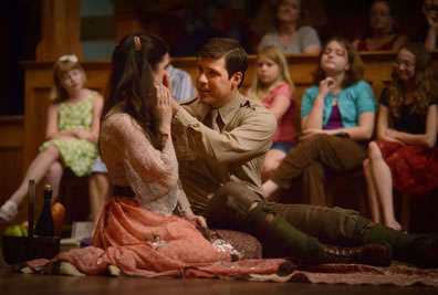 Bertram in mid-20th century british military uniform--khaki shirt and tie, brown pants, olive stockings, brown boots--sits on the floor and has his hands up to Diana's cheek as she has her right hand on his; she's in colorful print lacey blouse and peach dress.