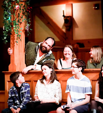 Benedick in green renaissance vest jacket over white shirt leans out from behind a vine-covered post next to two laughing women with three kids on the gallant stools in front of him, one boy looking up at him.