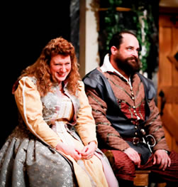 Beatrice in ornate yellow and silver dress, ands on her lap and snickering as she looks down to the right, next to her Benedick in black vest over rust and burgundy colored renaissance jacket and breeches, rolling his eyes looking up and to the left, both sitting on a bench