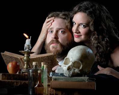 Mephistopheles with her check pressed against Faustus's temple, her right hand hugging his head, he's sitting at a desk with a book open, a candle, a skull on stacked books, an apple on another stack of books, and vials between the stacks