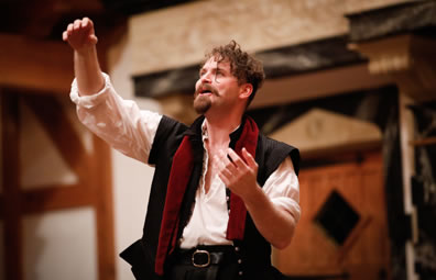 Hamlet looking up, motioning with both arms, wearing white shirt, black renaissance vest and  a red scarf draped over his shoulder