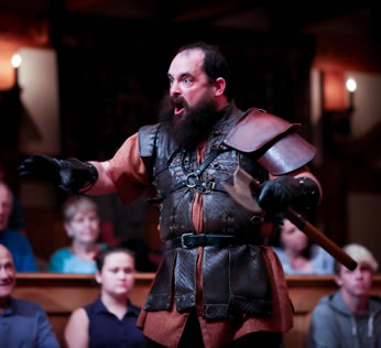 Cade in leath armor over a worker's tunic, black gloves, and a huge beard as he holds a stick and roars with eyes bulging. Audience sits in the background.