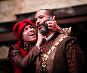 The Fool in red jesters cap, brown shirt, red vest, and colorful collar uses his fingers on Lear's bearded cheek to make him smile as Lear stares vacantly ahead, wearing regal shirt, brown leather vest, and a medallion on a chain.