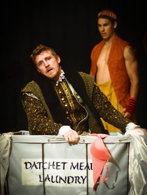 Ford in richly embroidered tudor cloak looking forlorn as he kneels in the laundry basket, which has a pink bra hanging over the front. Stenciled on the basked is "Datchet Mead Laundry." In the background is the servant, shirtless with orange vest and baggy yellow pants