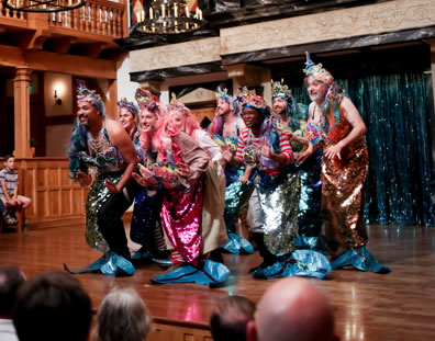 Nine mermaids, each in multi-colored metalic fins with blue flippers and various crowns, dance in a diamond on the center of the stage, with audience sitting on the stage behind and chandeliers of the Playhouse hanging overhead.