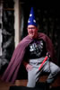 Arragon on one knee, blue star and moon wizard hat, brown-framed glasses, purple cape, "Choose Your Weapon" black t-shirt, gray sweatpants, and red light saber in his left hand, right hand on hip.