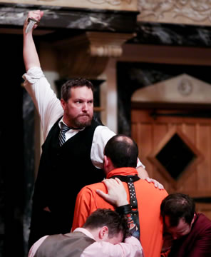Shylock in black vest, white shirt, and black and white-striped tie holds his knife aloft but looks off to the side toward Portia, his hand on the orange-prison-garb clad Antonio (back to us) as Bassanio and Gratziano, heads down, brace Antonio.