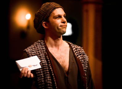 Truffaldino in checkered knit cap, rug-like stole and ragged t-shirt holds up an opened letter
