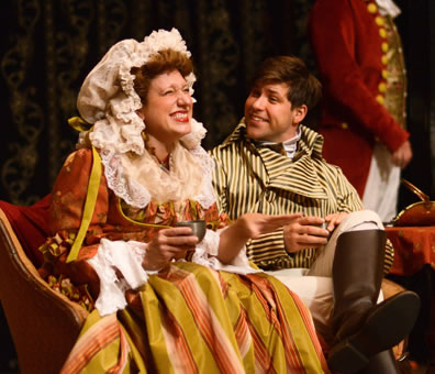 Mrs. Hardcastle in orange and gold striped dress and white matrons cap sits on a settee with Hastings in yellow and black striped coat, white pants and brown riding boots, both holding punch, the lady laughing and Hastings looking upon her bemused.