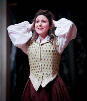Judith, in simple Elizabethan waist jacket, blousey shirt and maroon dress holds her hands up to her head in wonder.