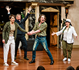 Petruchio in striped shirt and torn pants holds Katherina's left hand with both of his. He's shouting, she, in hooded sweater and sweat pants, is looking dumbfounded. Behind them, Baptista in a suit has his hands raised, to the right Tranio in panama hat and jacket has her arms outstretched, and to the left Gremeo is leaning on his cane.