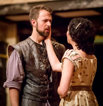 Leantio is wearing a leather renaissance  vest with buckles down the front and a purple shirt with rolled up sleeves. Bianca in simple tan dress with a pattern of brown brances and leaves holds her right hand on Leantio's bearded cheek.