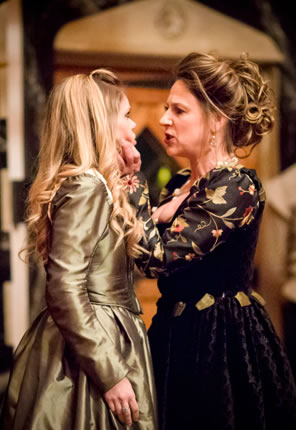 Livia snarling in the face of and her left hand gripping the chin of Isabella, who is wearing a classic greenish gold Elizabethan dress