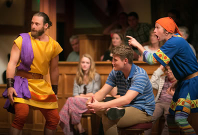 Antipholus with a beard looks to his right in a state of readiness, knees bent, while across from him Dromio points a finger and has his mouth open. Between them, a young man sits with right ankle on left knee, leaning forward with hands clasped, and wearing a blue and white striped polo shirt and brown pants. In the background is more audience. 