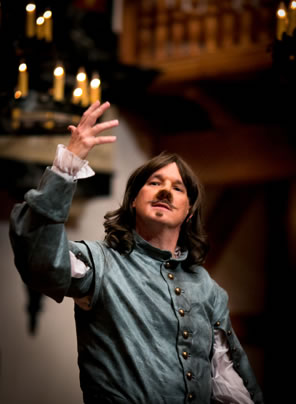 Cyrano with right hand upraised and wearing a blue jacket with frilly cuffed white shirt, shoulder-length hair