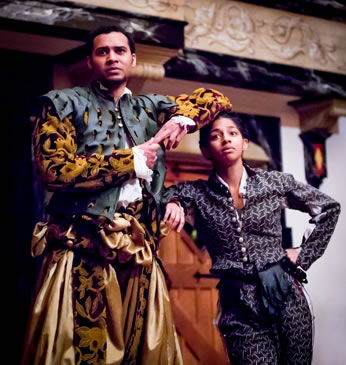 Mercutio in gold sattin pants with gold and black patterned felt jacket and drapery, cut leather black vest, big gold bows for a belt, and white fluffy shirt stands with his left arm perched on the head of Benvolio, who's leaning with his right arm on Mercutio's hilt and left fist on his own hip and wearing a blue-patterend pants and shirt with leath gloves hanging in his belt