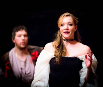 Angellica wearing a black strapless gown with white puffy sleeves, bare shoulders and a jewelled choker, her hair twirling down one side of her neck, is facing the camera, holding he rleft hand up, and out of focus in the background is Willmore, white tunic, red ribbons on his brown vest, looking achingly at her