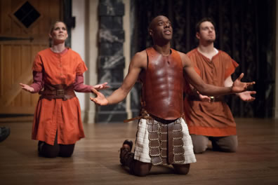 Palamon in leather breast plat and skirt kneels with arms spread, head back and eyes closed, behind him in orange leather skirts are his companion knights
