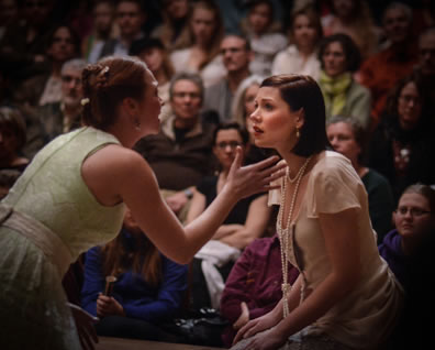 Celia in green dress talks demonstravely with Rosalind in tan dress and pearls with the theater audience in the backgroun