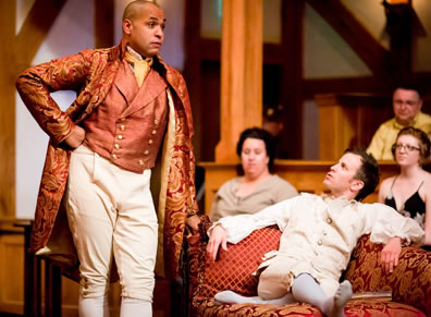 Scandal in dark peach vest, orange cravat, orange embroidered cloak, cream knee britches with right first on hip and left hand leaning on chaiz lounge on which lies Valentine in white puffy shirt and kneebritches and in white stockinged feet, with audience in background
