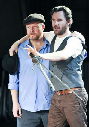 Hamlet in vest and brown jeans holding a sword and dagger with his arms around Horatio in Irish cap and blue shirt, a bowler gripped in Hamlet's other hand.