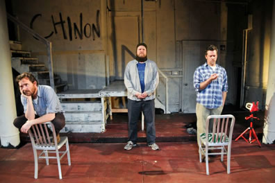 Tucker squating on a metal chair, Edmunds standing in the center, and O'Keefe standing behind another metal chair with "France" painted in green on the seat, and on the wall in the background is painted "Chinon"