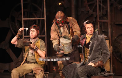 Dromio in old leather flight cap, tan work shirt and pants with vest holds up a spoon dripping white gunk into a bowl in his left hand, a man in industrial clothes, leather helmet and red goggles pours slime into another bowl from an old metal watering can, the bowl is on a table made of a metal cog on wood stand, and Antipholus sits wearing long gray overcoat, gold vest and cravat. The chairs have ladders for backs, and in the background are gears, tubes and valves on the wall