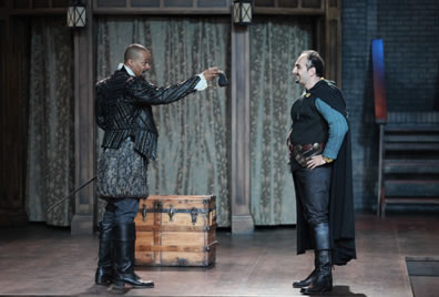 Burbage in Elizabethan leather jacket, bulging knee britches and boots holds out a black purse to Shakespeare in jacket vest over blue shirt and black pants with boots and long cloak. A wood trunk is on the stage.