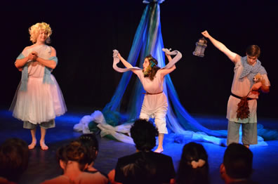 Flute as Thisbe wears a ballerina dress over his shin-length pants and bare feet, a blue scarf draped around his arms, and a blond wig with hands clasped together; Snug as Lion has her hands raised holding the mantle across the back of her neck, flowers in her hair; Starveling as Moonshine holds up the lantern in his right hand, holds a stuffed dog in the crook of his left arm and holding a thorn bush in that hand, his head down.  We see the backs of the noble audience's heads. Maypole is in the background.