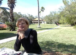 Katrina Boler, in dark gray sweater, leans on her elbow at a picnic table with The Barnacle house and lawn in the background and people walking on a path