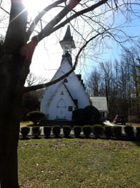 The church, white with gray shingle roof, a capped steeple at the front, trees all around, an extension jutting out at the back right.