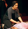 Antonia, in black blouse with black and white striped shoulders and sleeves to elbows and a bow on the collar and black pants kneels next to Caesar's bloody body; she's holding a knife in her righ hand and looking up at someone beyond the body
